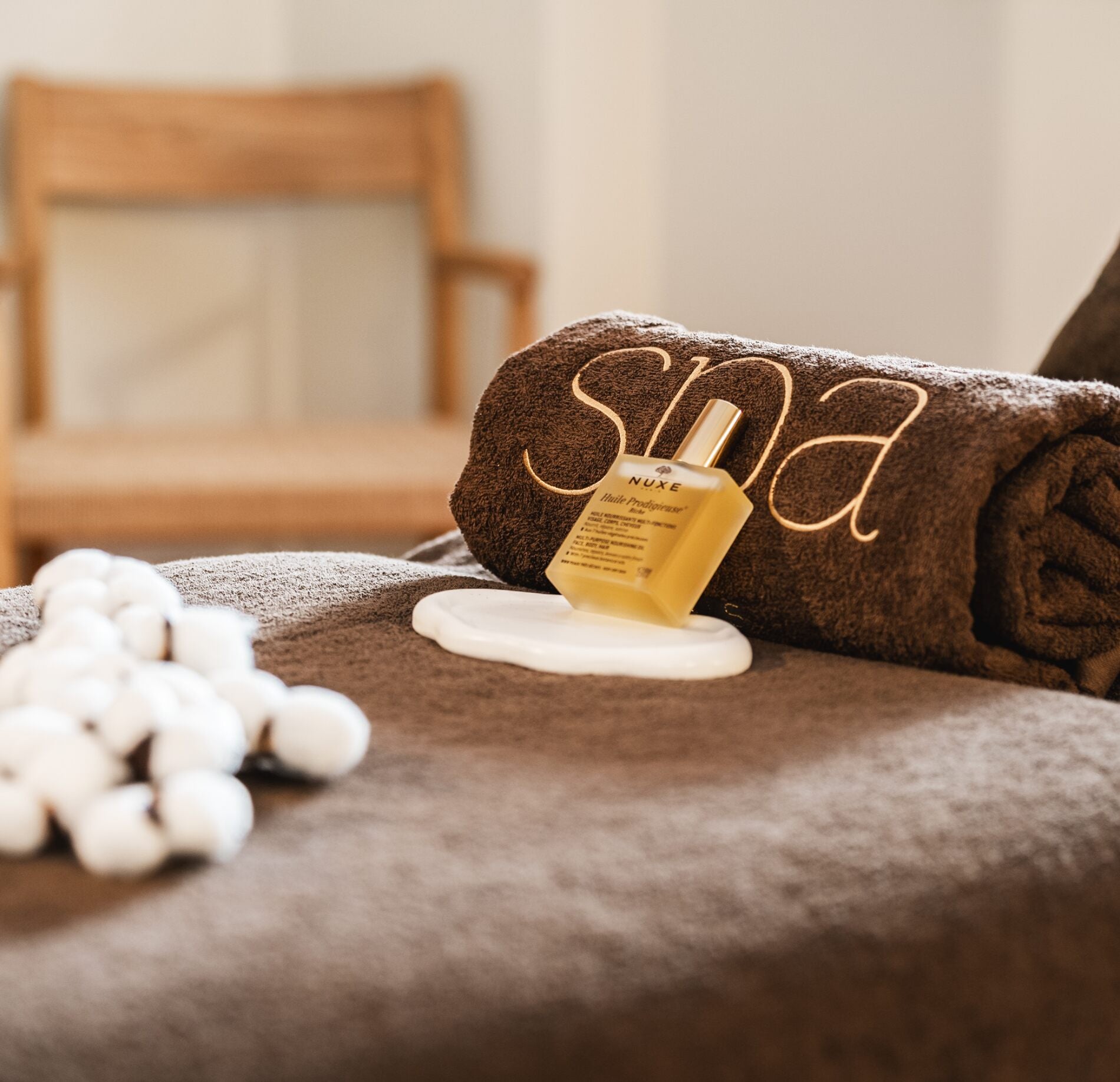 Un détail de soin raffiné du Spa la Villa Camille, avec une serviette brune roulée portant l’inscription “spa” et un flacon de soin posé délicatement par-dessus.
Au premier plan, des accessoires blancs et une texture douce créent une ambiance chaleureuse, tandis qu’un fauteuil en bois flou en arrière-plan renforce l’atmosphère apaisante.