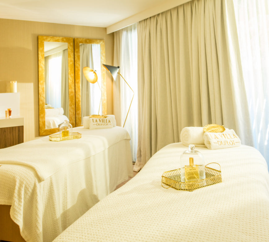 Une cabine de soin lumineuse du Spa Villa Duflot, avec deux tables de massage recouvertes de linge clair et surmontées de serviettes parfaitement pliées.
Le décor élégant, composé de rideaux beiges, de miroirs dorés et de plateaux avec flacons, diffuse une atmosphère chaleureuse et raffinée propice à la détente.