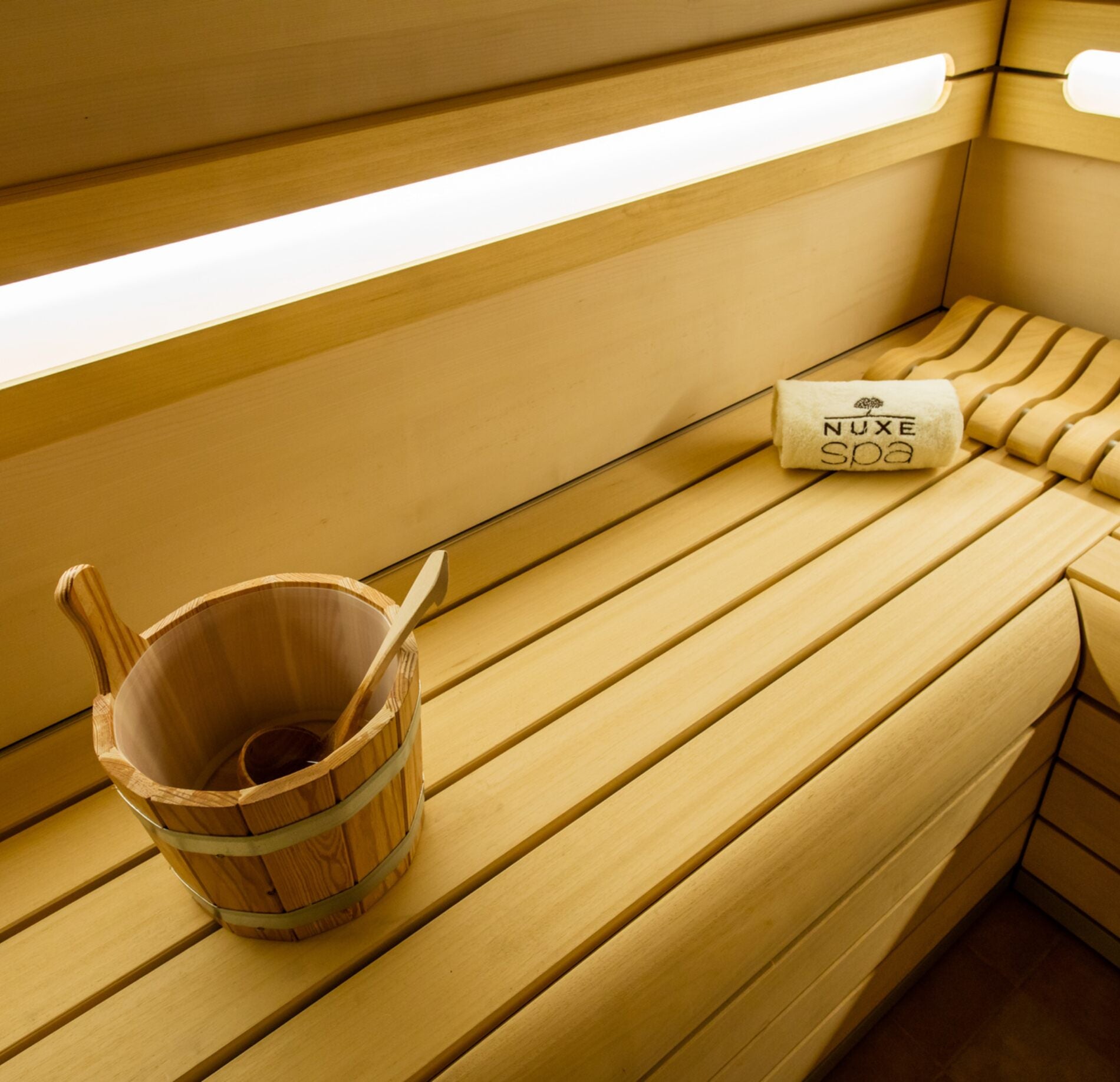 Un sauna en bois lumineux du Spa Villa Duflot, avec des bancs en lattes claires, un seau traditionnel en bois et une louche posée sur le côté.
L’éclairage linéaire intégré au mur diffuse une lumière douce et chaleureuse, mettant en valeur les textures naturelles et l’ambiance apaisante de l’espace.