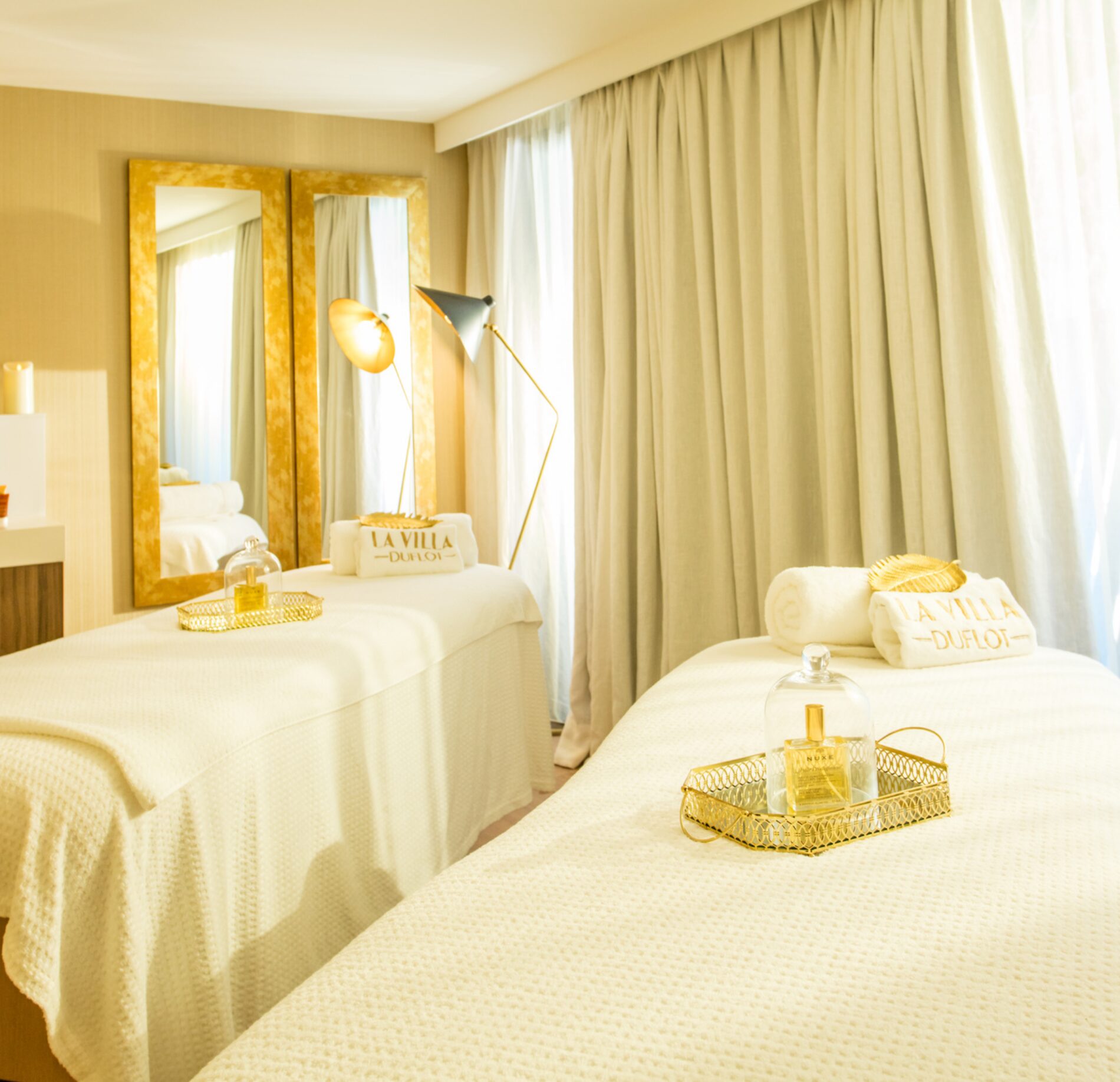 Une cabine de soin lumineuse du Spa Villa Duflot, avec deux tables de massage recouvertes de linge clair et accompagnées de serviettes soigneusement pliées.
Le décor raffiné, composé de rideaux beiges, de miroirs dorés et de plateaux avec flacons, crée une atmosphère chaleureuse et apaisante propice à la détente.