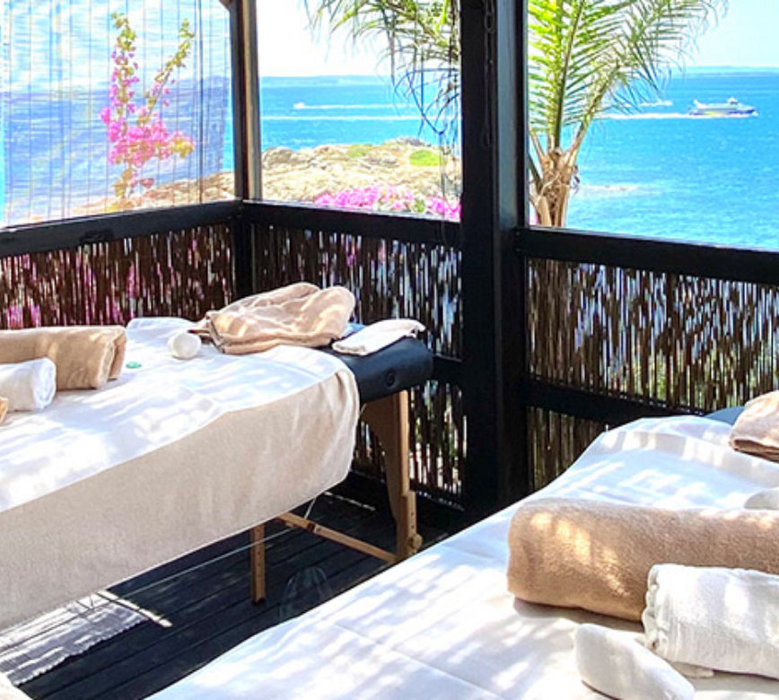 Une cabine de soin en plein air du Spa Hotel Vistabella, avec deux tables de massage recouvertes de linge clair et disposées face à la mer.
À travers une structure en bois ajourée, la vue s’ouvre sur une eau bleu intense et une végétation fleurie, créant une atmosphère lumineuse et apaisante.