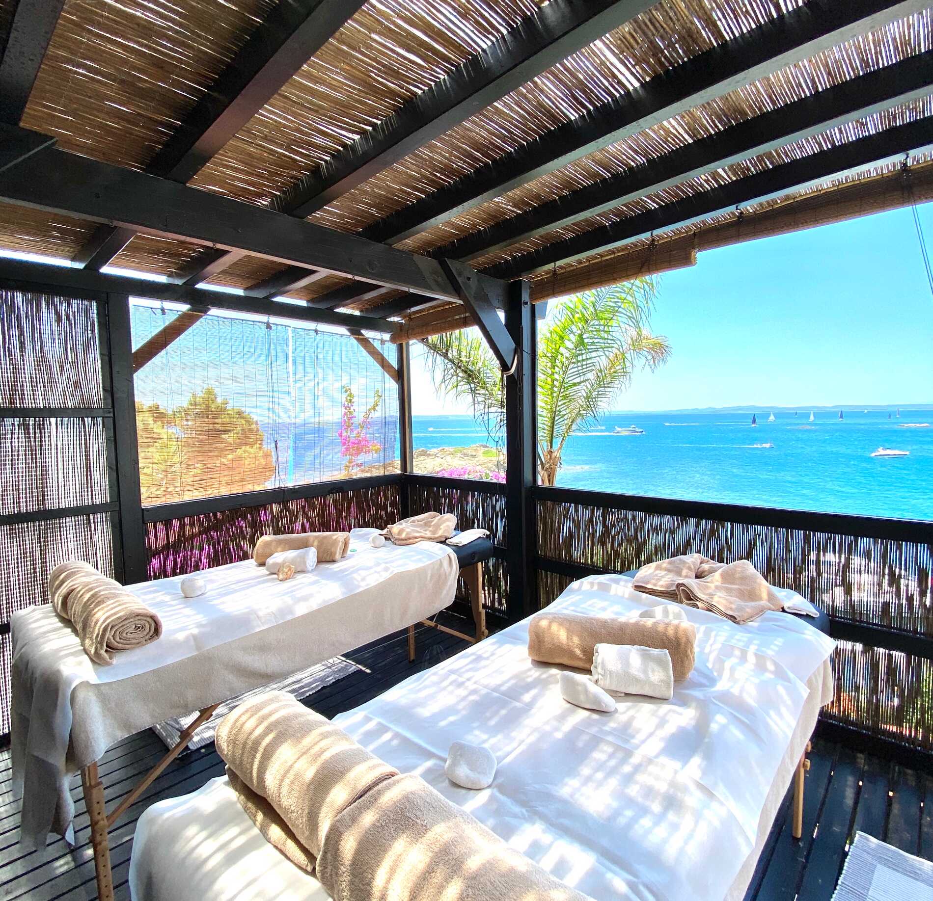 Une cabine de soin en plein air du Spa Hotel Vistabella, avec deux tables de massage recouvertes de linge clair et installées face à la mer.
À travers une structure en bois ajourée, la vue s’ouvre sur une eau bleu profond et une végétation fleurie, créant une atmosphère lumineuse et relaxante.