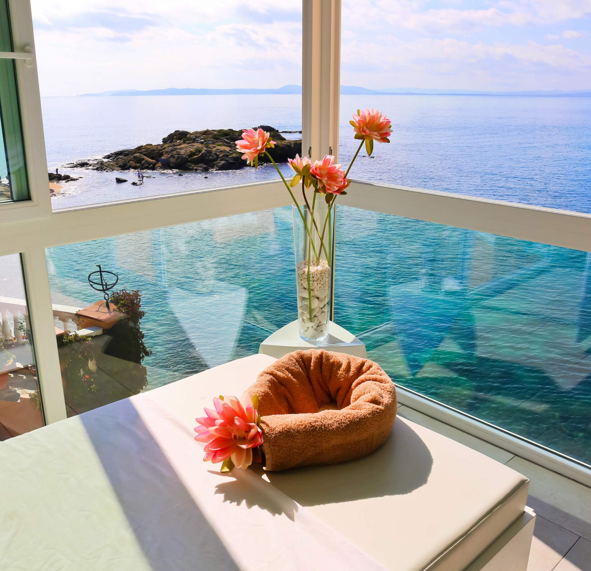 Une cabine de soin lumineuse du Spa Hotel Vistabella, avec une table de massage installée face à une baie vitrée offrant une vue panoramique sur la mer.
Au premier plan, une serviette roulée et un vase de fleurs colorées apportent une touche délicate, tandis que l’eau turquoise et les rochers créent une atmosphère apaisante et ouverte.