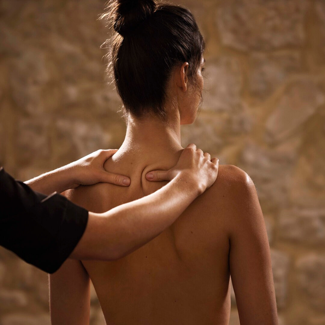 Une personne, vue de dos avec les cheveux attachés, reçoit un massage au niveau de la nuque et des épaules. Les mains du praticien exercent une pression précise de part et d’autre du cou. L’ambiance est chaleureuse, avec un fond en pierre qui évoque un espace de spa apaisant.