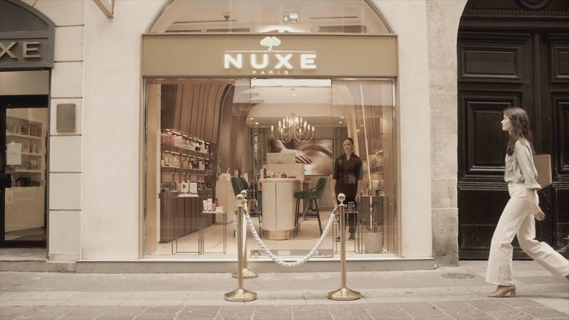 On découvre une femme élégante qui pousse la porte d’une boutique NUXE à Paris. Elle entre avec curiosité et raffinement, prenant le temps d’observer les différentes gammes de la marque exposées sur les étagères. Les produits, variés et soigneusement présentés, attirent son regard tandis qu’elle avance doucement dans cet univers dédié au bien‑être et à la beauté.
Attirée par l’espace spa, elle s’y dirige naturellement. Elle est alors accueillie chaleureusement par une spécialiste, qui l’invite à s’installer